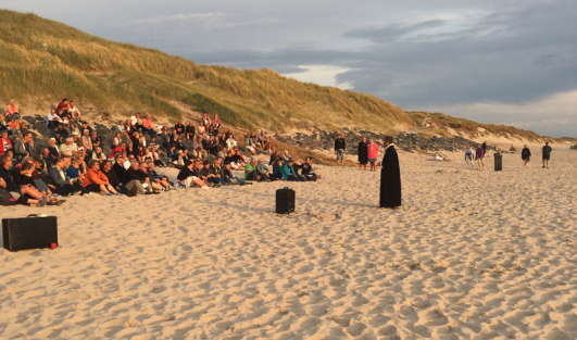 Forsamling sidder på skrænt ved stranden og kigger på præst
